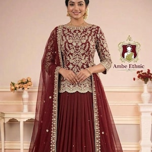 Puede incluir: Un vestido étnico granate con intrincados bordados dorados. El vestido presenta un corpiño ajustado con mangas tres cuartos y una falda acampanada. Un dupatta transparente a juego con ribete dorado completa el conjunto. El logotipo de Ambe Ethnic es visible.