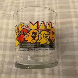 May include: A clear glass tumbler featuring the Pac-Man logo and characters in yellow, red, and orange. The text "PAC-MAN" is in yellow and red. The bottom of the glass has a thick, rounded base. The copyright information is visible.