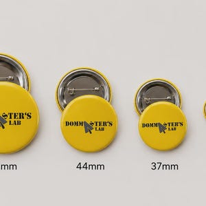 Può includere: Quattro spille rotonde gialle con un'icona a cursore nera e il testo "DOMMSTER'S LAB" stampato sul davanti. Le spille sono disposte per dimensione, etichettate come 58mm, 44mm, 37mm e 25mm, mostrando il meccanismo a spilla in metallo sul retro.