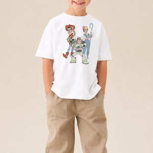 Puede incluir: Una camiseta blanca de manga corta con un estampado gr&aacute;fico colorido de tres personajes animados: un vaquero con sombrero, un guardi&aacute;n espacial con traje blanco y verde, y una pastora con un bast&oacute;n azul en la zona del pecho.