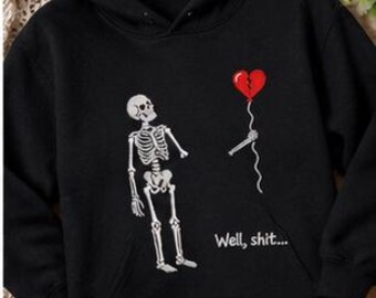 Könnte beinhalten: Ein schwarzer Kapuzenpullover mit einer Grafik eines weißen Skeletts, das auf einen roten, zerbrochenen Herzballon blickt, der von einer abgetrennten Skeletthand gehalten wird. Der Text "Well, shit..." ist in Weiß unter dem Ballon gedruckt.