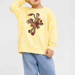 Puede incluir: Una sudadera de manga larga de color amarillo p&aacute;lido con un estampado gr&aacute;fico de un vaquero de dibujos animados montando un caballo marr&oacute;n. El vaquero lleva una camisa amarilla, un chaleco azul y un pa&ntilde;uelo rojo, mientras sostiene un sombrero.