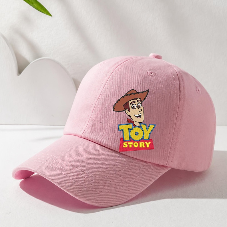 Puede incluir: Una gorra de b&eacute;isbol de color rosa claro con un estampado gr&aacute;fico del personaje Woody de Toy Story. El dise&ntilde;o incluye la cabeza del personaje con un sombrero de vaquero marr&oacute;n y un pa&ntilde;uelo rojo, sobre el logotipo amarillo y azul de Toy Story.
