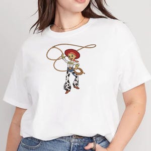 Puede incluir: Una camiseta blanca de manga corta con un estampado gr&aacute;fico de un personaje de vaquera de dibujos animados con pelo rojo, sombrero rojo y pantalones con estampado de manchas de vaca, sosteniendo un lazo marr&oacute;n sobre su cabeza.