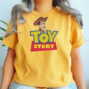 Puede incluir: Una camiseta de color amarillo mostaza con un bordado del personaje Woody de Toy Story. El dise&ntilde;o incluye el sombrero de vaquero marr&oacute;n, el rostro del personaje y el logotipo azul y amarillo de Toy Story sobre una base rectangular roja.