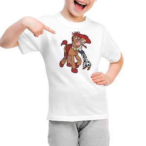 Puede incluir: Una camiseta blanca de manga corta con un estampado gr&aacute;fico de un caballo marr&oacute;n con una silla de montar roja y una vaquera de pelo rojo, sombrero rojo y pantalones con manchas de vaca abrazando el cuello del caballo con cari&ntilde;o.