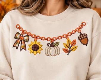 Könnte beinhalten: Ein beigefarbenes Sweatshirt mit Rundhalsausschnitt und einer aufgestickten orangefarbenen Kette auf der Brust. An der Kette hängen fünf herbstliche Anhänger: eine karierte Schleife, eine gelbe Sonnenblume, ein weißer Kürbis, bunte Blätter und eine Eichel.