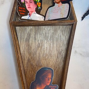 May include: A coffin-shaped wooden shelf with three compartments, decorated with stickers. The stickers feature portraits with text: "does he feel like home", "Just another sacrifice", and "Ghosts are real. This much I know". A small mushroom figurine is at the bottom.