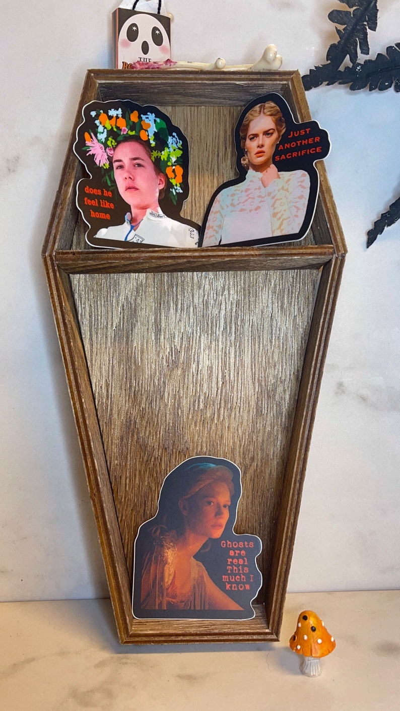 May include: A coffin-shaped wooden shelf with three compartments, decorated with stickers. The stickers feature portraits with text: "does he feel like home", "Just another sacrifice", and "Ghosts are real. This much I know". A small mushroom figurine is at the bottom.