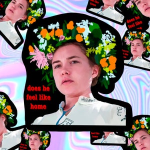 May include: A digital art print featuring a woman with a floral crown and the text "does he feel like home" in red. The image has a black border and is set against a colorful, iridescent background. The woman is wearing a white top.