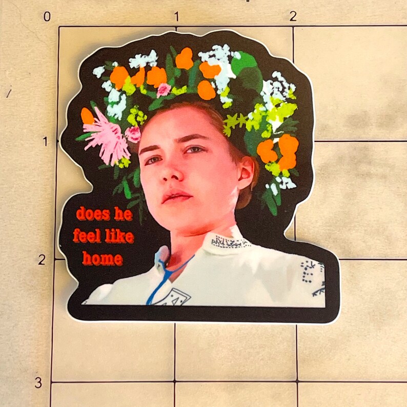 May include: A sticker featuring a woman with a floral crown and the text "does he feel like home" in red. The sticker has a black border and a white background. The woman is wearing a white top.
