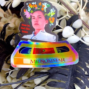 May include: Two stickers featuring imagery from the film Midsommar. One sticker depicts a woman with a floral crown, with the text "does he feel like home." The other is a holographic cassette tape with the film's title.