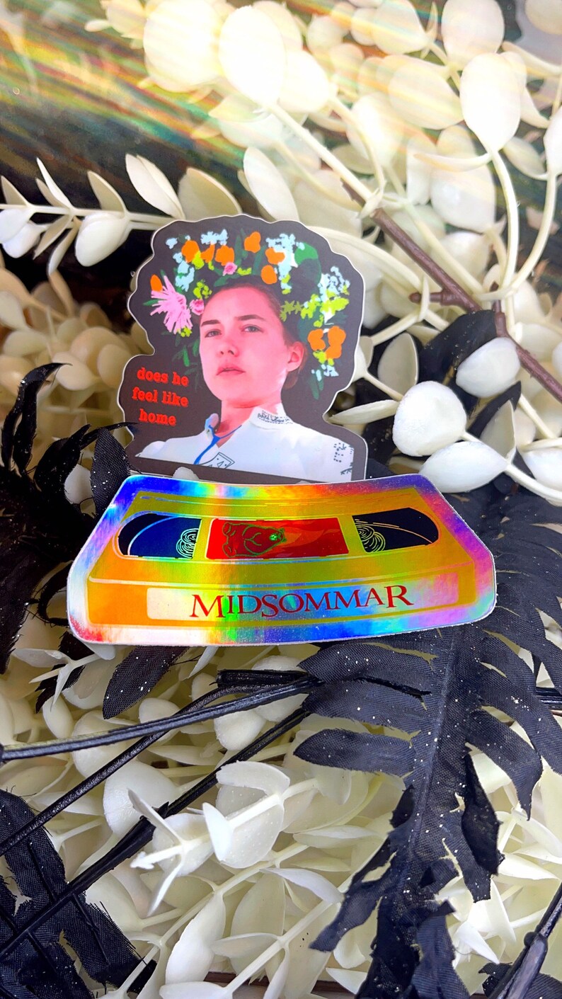 May include: Two stickers featuring imagery from the film Midsommar. One sticker depicts a woman with a floral crown, with the text "does he feel like home." The other is a holographic cassette tape with the film's title.