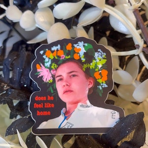 May include: A sticker featuring a woman with a floral crown and the text "does he feel like home." The sticker has a black border and is set against a backdrop of white and black artificial foliage.