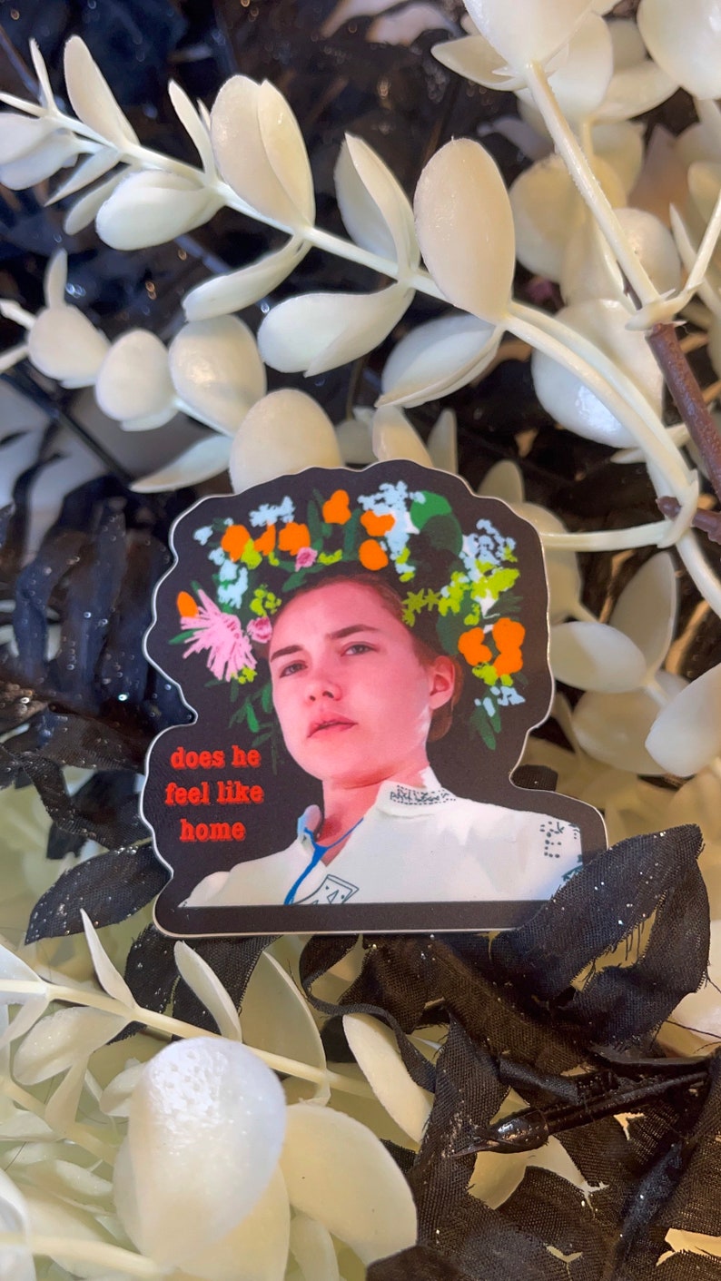 May include: A sticker featuring a woman with a floral crown and the text "does he feel like home." The sticker has a black border and is set against a backdrop of white and black artificial foliage.