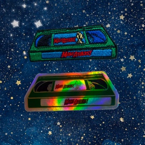 May include: Two patches featuring a VHS tape design with the text "Mars Attacks!". The top patch is embroidered with green, blue, and black thread. The bottom patch is a holographic sticker with a green border and the same text.