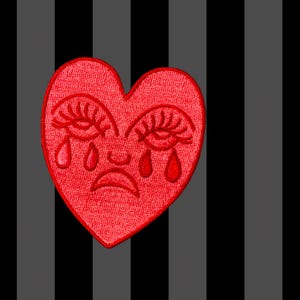 May include: A red heart-shaped embroidered patch with a sad face design. The heart has crying eyes and teardrops. The background features vertical stripes in black, grey, and dark grey.