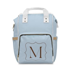 Puede incluir: Una mochila azul claro con ribetes blancos y un asa blanca. El bolsillo delantero presenta un marco decorativo marrón con la letra "M". La mochila tiene cremalleras doradas y bolsillos laterales.