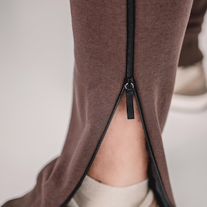 Op de afbeelding: Close-up van een bruine joggingbroek met een zwart ritsdetail langs de buitennaad van het onderbeen. De rits is gedeeltelijk open, waardoor een glimp van een beige sneaker en enkel te zien is. De broek is gemaakt van een zachte, getextureerde stof.