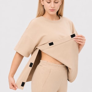 Op de afbeelding: Een beige top met korte mouwen en een ronde hals, voorzien van een zijopening die wordt vastgezet met zwarte klittenbandsluitingen. De top is gecombineerd met een bijpassende beige broek met een hoge tailleband en zijzakken.