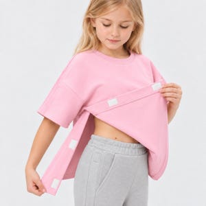 Op de afbeelding: Een lichtroze sweatshirt met korte mouwen, een ronde hals en zijsplitten die zijn vastgezet met witte klittenbandsluitingen, gecombineerd met een lichtgrijze joggingbroek met een hoge tailleband en zichtbare zijzakken aan de voorzijde.