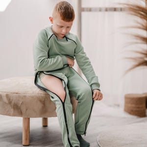 Puede incluir: Un conjunto verde claro de sudadera y pantalón con detalles de cremallera negra. Los pantalones tienen cremalleras que van desde la cintura hasta los tobillos. El conjunto se usa con calcetines verdes a juego. La ropa está diseñada para la comodidad y el estilo.