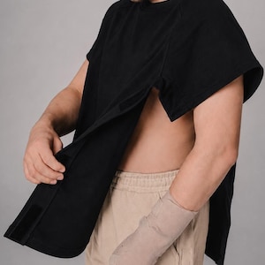 Op de afbeelding: Een zwart shirt met korte mouwen en een zijopening die is vastgezet met klittenband, gecombineerd met een beige korte broek. Het shirt heeft een losse pasvorm en een ronde hals, ontworpen om gemakkelijke toegang tot de romp te bieden.