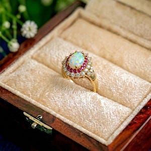 Könnte beinhalten: Ein goldener Ring mit einem ovalen Opal-Mittelstein, umgeben von kleinen roten Edelsteinen und klaren Akzentsteinen. Der Ring ist in einem goldenen Band gefasst und wird in einer samtgefütterten Holzschatulle präsentiert.