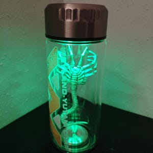 Op de afbeelding: Een heldere glazen waterfles met een roestvrijstalen deksel en basis. Binnenin is een groen oplichtend Xenomorph-figuur zichtbaar. De fles heeft een groen licht en de tekst "WE-AND-YU-TAM CORP" staat op de zijkant.