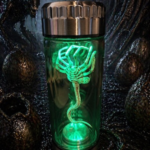 Può includere: Un contenitore cilindrico in vetro con tappo e base in metallo argentato, dotato di una luce verde interna che illumina una creatura pallida dalle molteplici zampe con una lunga coda segmentata sospesa all'interno del recipiente.