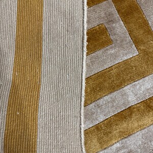 Puede incluir: Una alfombra con un patr&oacute;n geom&eacute;trico de rayas amarillo mostaza y beige. El lado izquierdo muestra el reverso con una estructura tejida texturizada, mientras que el derecho exhibe la superficie de pelo cortado con un dise&ntilde;o angular.