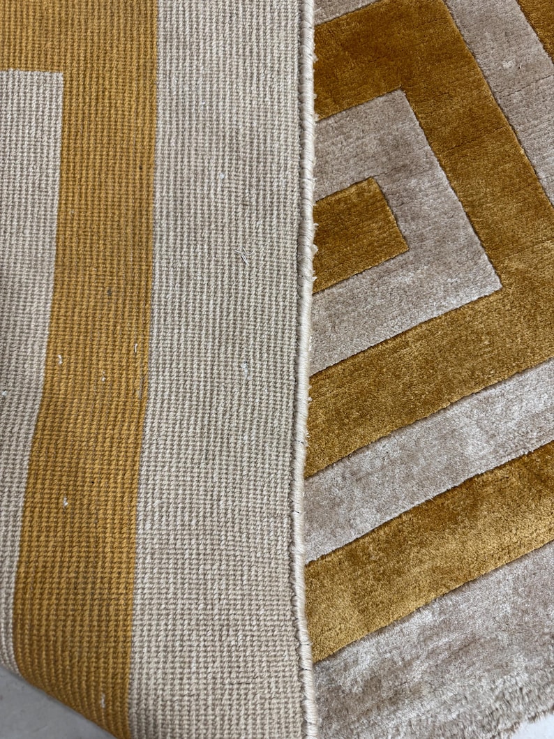 Puede incluir: Una alfombra con un patr&oacute;n geom&eacute;trico de rayas amarillo mostaza y beige. El lado izquierdo muestra el reverso con una estructura tejida texturizada, mientras que el derecho exhibe la superficie de pelo cortado con un dise&ntilde;o angular.