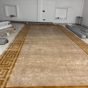 Puede incluir: Una alfombra rectangular grande con un centro beige s&oacute;lido y un borde ancho decorado con un patr&oacute;n de greca griega dorada repetitivo. La alfombra tiene una textura lisa de pelo corto y est&aacute; extendida sobre el suelo en una habitaci&oacute;n amplia.