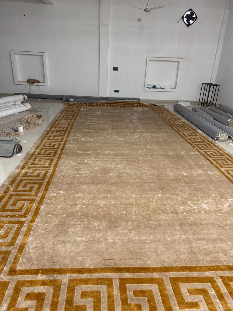 Puede incluir: Una alfombra rectangular grande con un centro beige s&oacute;lido y un borde ancho decorado con un patr&oacute;n de greca griega dorada repetitivo. La alfombra tiene una textura lisa de pelo corto y est&aacute; extendida sobre el suelo en una habitaci&oacute;n amplia.