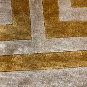 Puede incluir: Una alfombra rectangular con un patr&oacute;n geom&eacute;trico de bandas alternas en color amarillo mostaza y beige claro. La alfombra presenta una textura densa de pelo corto con l&iacute;neas horizontales visibles y un aspecto suave y afelpado.