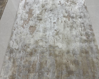 Puede incluir: Una alfombra rectangular con un patrón desgastado y moteado en tonos plata, gris claro y beige apagado. La superficie tiene una apariencia texturizada e irregular con alturas de pelo variables que crean un efecto visual abstracto.