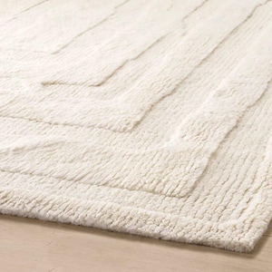 May include: Cream-colored, textured rug with a geometric pattern. The rug features a series of raised, rectangular shapes arranged in a zig-zag design. The rug's plush texture and neutral color make it suitable for various interior styles.
