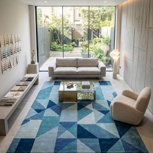 Peut inclure: Salon moderne avec un grand tapis géométrique bleu. Un canapé beige, une table basse dorée et en verre, et un fauteuil crème sont disposés sur le tapis. De grandes fenêtres donnent sur un jardin, créant un espace lumineux.