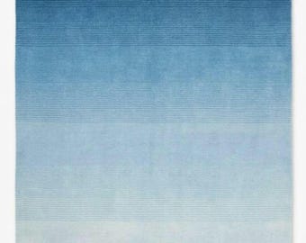 Puede incluir: Una alfombra rectangular con un patrón de degradado horizontal que transita desde un azul marino profundo en la parte superior hasta un azul cielo pálido y claro en la parte inferior. La superficie muestra líneas horizontales finas.