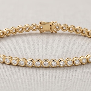 Puede incluir: Una pulsera de tenis dorada que presenta una hilera continua de piedras redondas, transparentes y facetadas, engastadas en biseles circulares individuales. La pulsera se asegura con un cierre de caja rectangular a juego con el tono dorado.