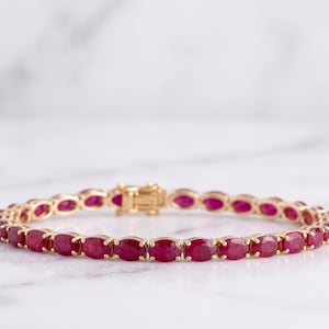 Könnte beinhalten: Ein goldenes Armband mit oval geschliffenen, tiefroten Edelsteinen. Die Edelsteine sind eng aneinander gesetzt und bilden eine durchgehende Linie. Das Armband hat einen sicheren Verschluss und liegt auf einer weißen Marmoroberfläche.