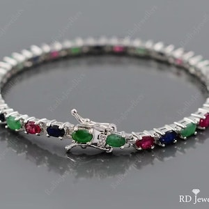 Könnte beinhalten: Ein silberfarbenes Tennisarmband mit einem sich wiederholenden Muster aus oval geschliffenen grünen, roten und dunkelblauen Edelsteinen in Metallfassungen. Das Armband verfügt über einen sicheren Kastenschloss-Verschluss mit Sicherheitsbügel.
