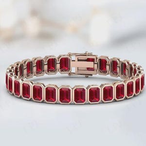 Könnte beinhalten: Ein roségoldenes Armband mit rechteckigen, tiefroten Edelsteinen. Das Armband besteht aus mehreren mit Edelsteinen besetzten Gliedern, die durch einen Verschluss verbunden sind. Die Edelsteine sind in einer Zargenfassung gefasst.
