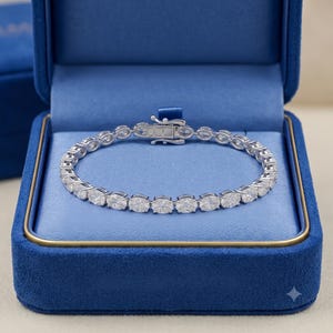 Puede incluir: Una pulsera de tenis plateada con una hilera continua de piedras transparentes de corte ovalado engastadas en metal. La pulsera incluye un cierre de caja seguro y se muestra dentro de un joyero forrado de terciopelo azul brillante.