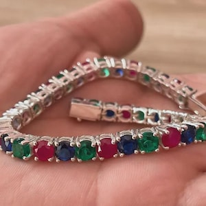 Könnte beinhalten: Ein silberfarbenes Tennisarmband mit einem sich wiederholenden Muster aus rund geschliffenen, in Krappen gefassten Edelsteinen in leuchtendem Rot, Blau und Grün. Das Armband besteht aus flexiblen Gliedern und einem sicheren Kastenschloss.