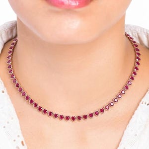 Könnte beinhalten: Eine Halskette mit einer durchgehenden Reihe kleiner, herzförmiger roter Edelsteine in einer Metallfassung. Die Steine sind in Größe und Form einheitlich und zu einer flexiblen Kette verbunden, die um den Halsansatz herum aufliegt.