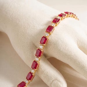 Könnte beinhalten: Ein goldenes Tennisarmband mit einem sich wiederholenden Muster aus rechteckigen, tiefroten Rubinen, die sich mit kleinen, runden, klaren weißen Diamanten abwechseln. Die Fassung ist aus Gelbgold und die Steine sind in flexiblen Gliedern gefasst.