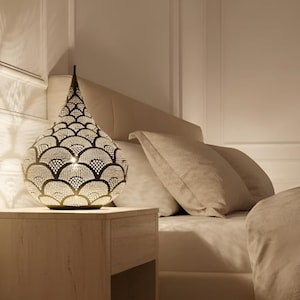 May include: A decorative, teardrop-shaped table lamp with a detailed, openwork metal design. The lamp is lit, casting a warm glow, and sits on a light-coloured nightstand next to a bed with neutral-toned bedding. The lamp's height is approximately 46 cm.