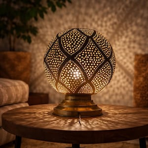Op de afbeelding: Decoratieve, bolvormige tafellamp met een geperforeerde metalen kap. De lamp straalt een warm, goudkleurig licht uit en werpt ingewikkelde schaduwpatronen op de houten tafel. De voet heeft een bijpassende goudkleur.