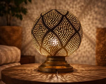May include: A decorative, spherical table lamp with a perforated metal shade. The lamp has a warm, golden glow, casting intricate patterns of light and shadow on the wooden table. The base is a matching gold color.